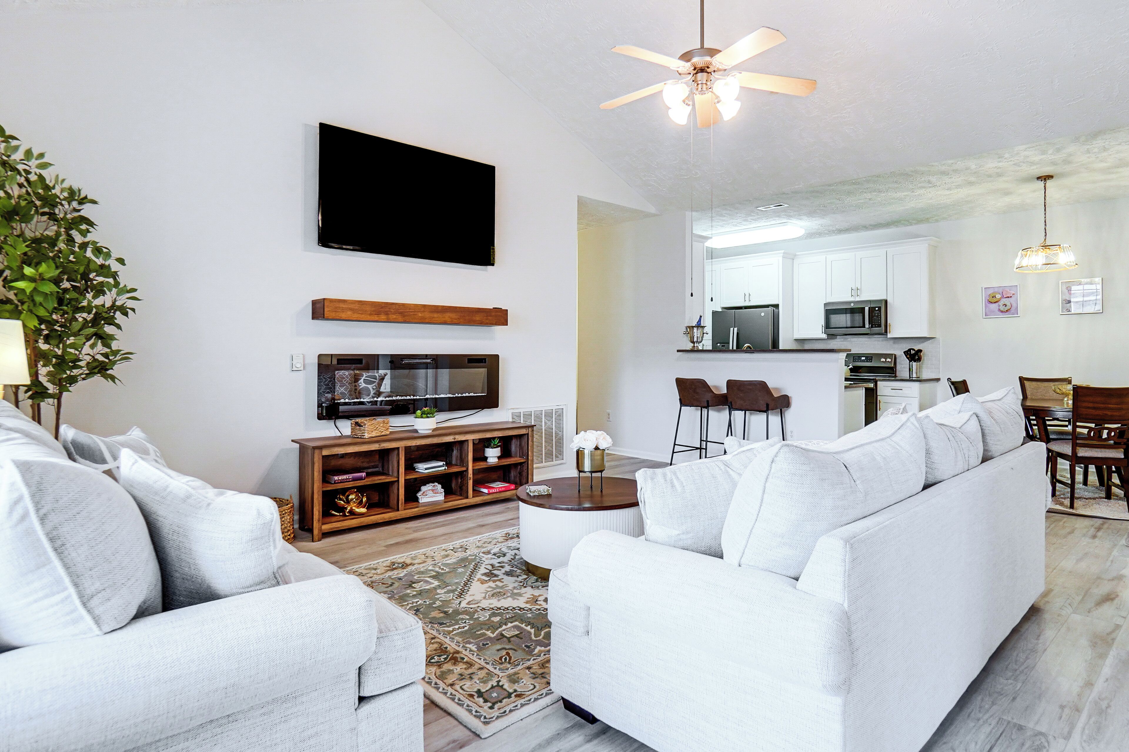 A modern living room with a flat-screen TV, a ceiling fan, and a dining area with a kitchen in the background.