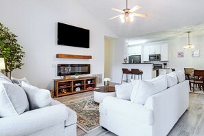 A modern living room with a flat-screen TV, a ceiling fan, and a dining area with a kitchen in the background.