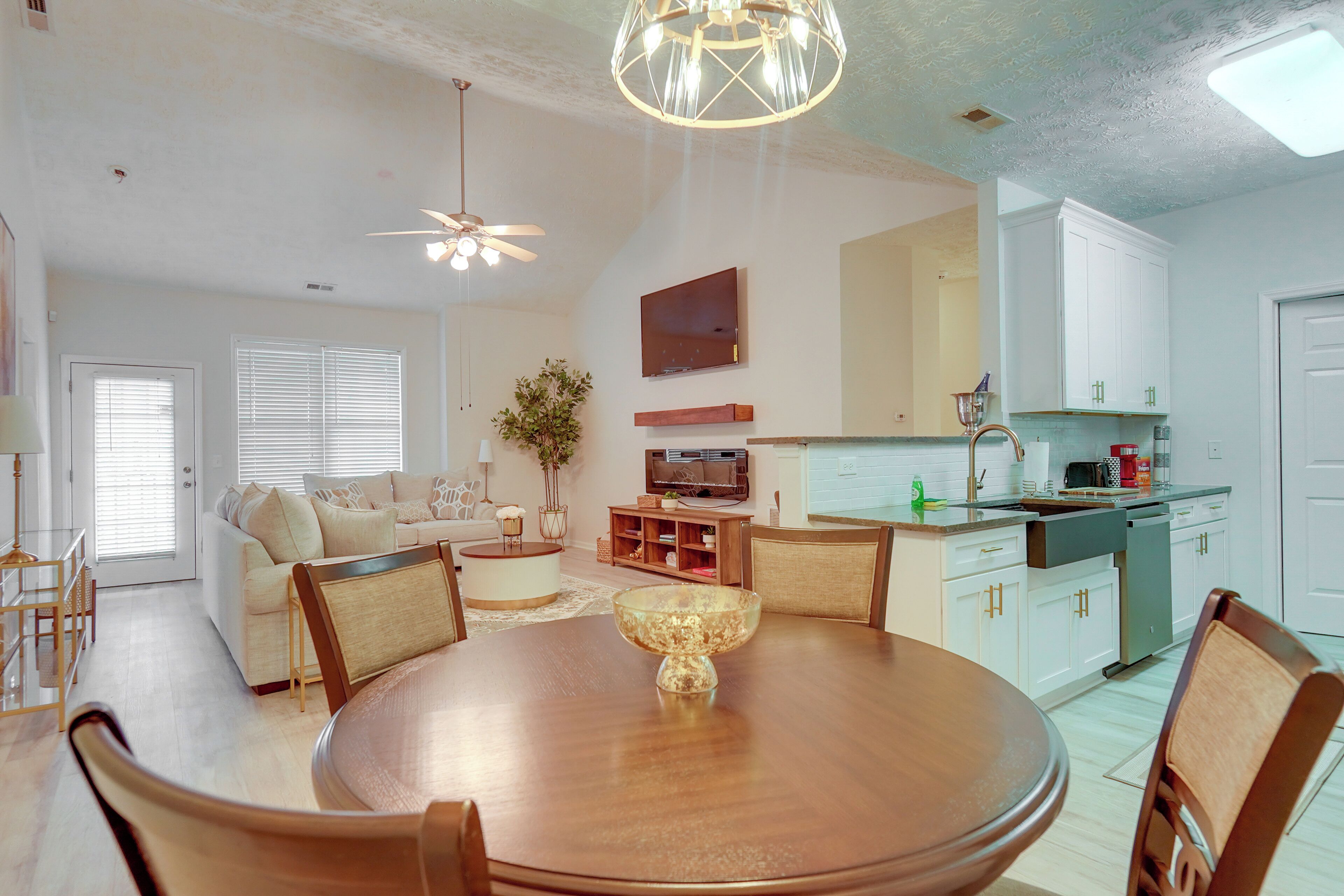 A modern kitchen with white cabinets, a dining area with a round table and chairs, and a living room with a sofa and a TV.