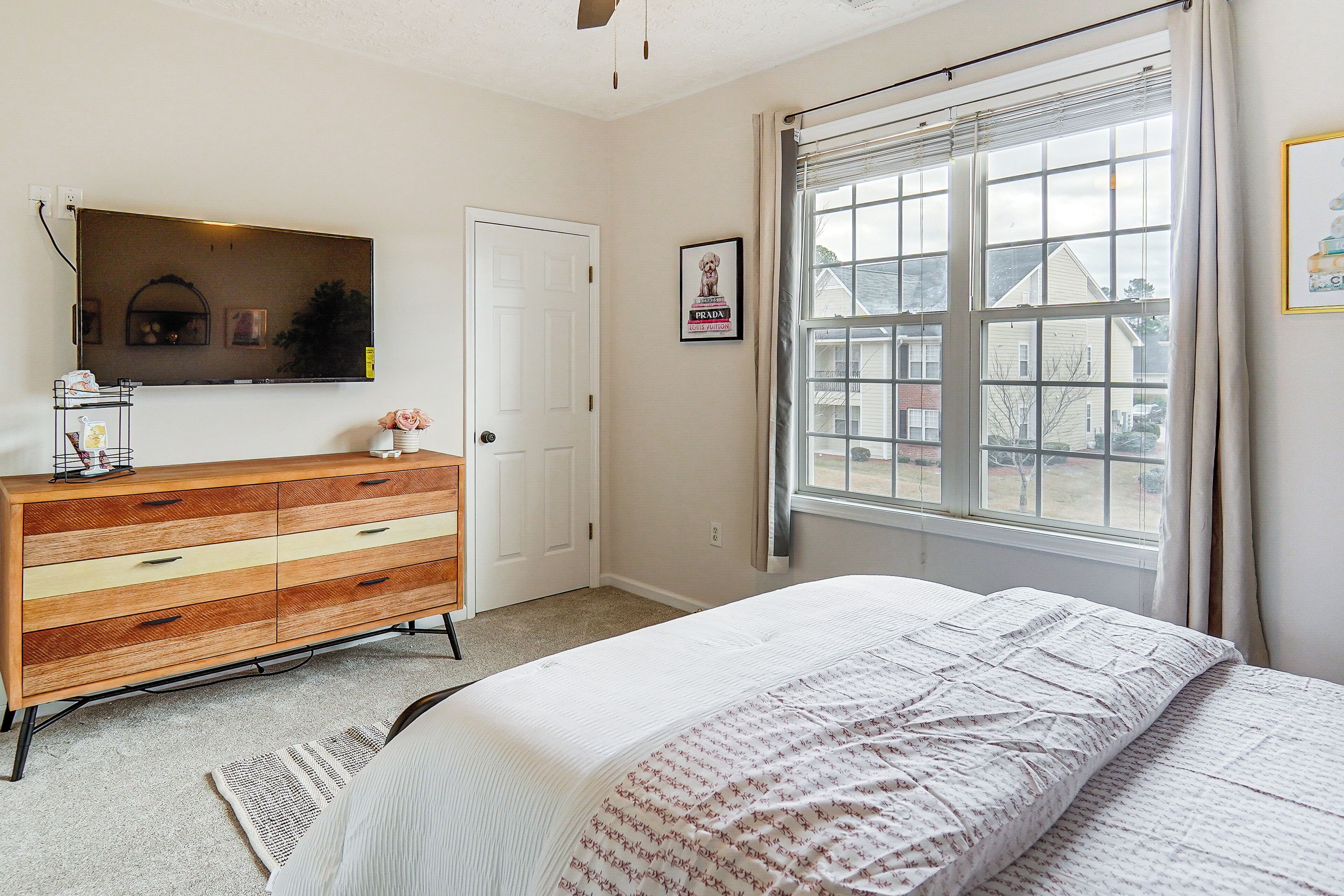 Une chambre à coucher avec un lit, une commode, une fenêtre avec des rideaux et un tableau encadré au mur.