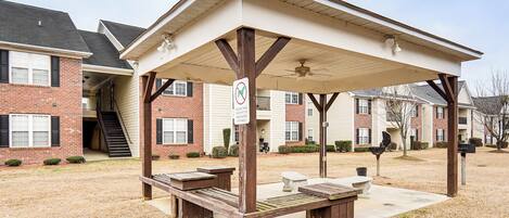 A covered outdoor seating area with picnic tables and benches, a no smoking sign, and a building with brick and siding exteriors in the background.