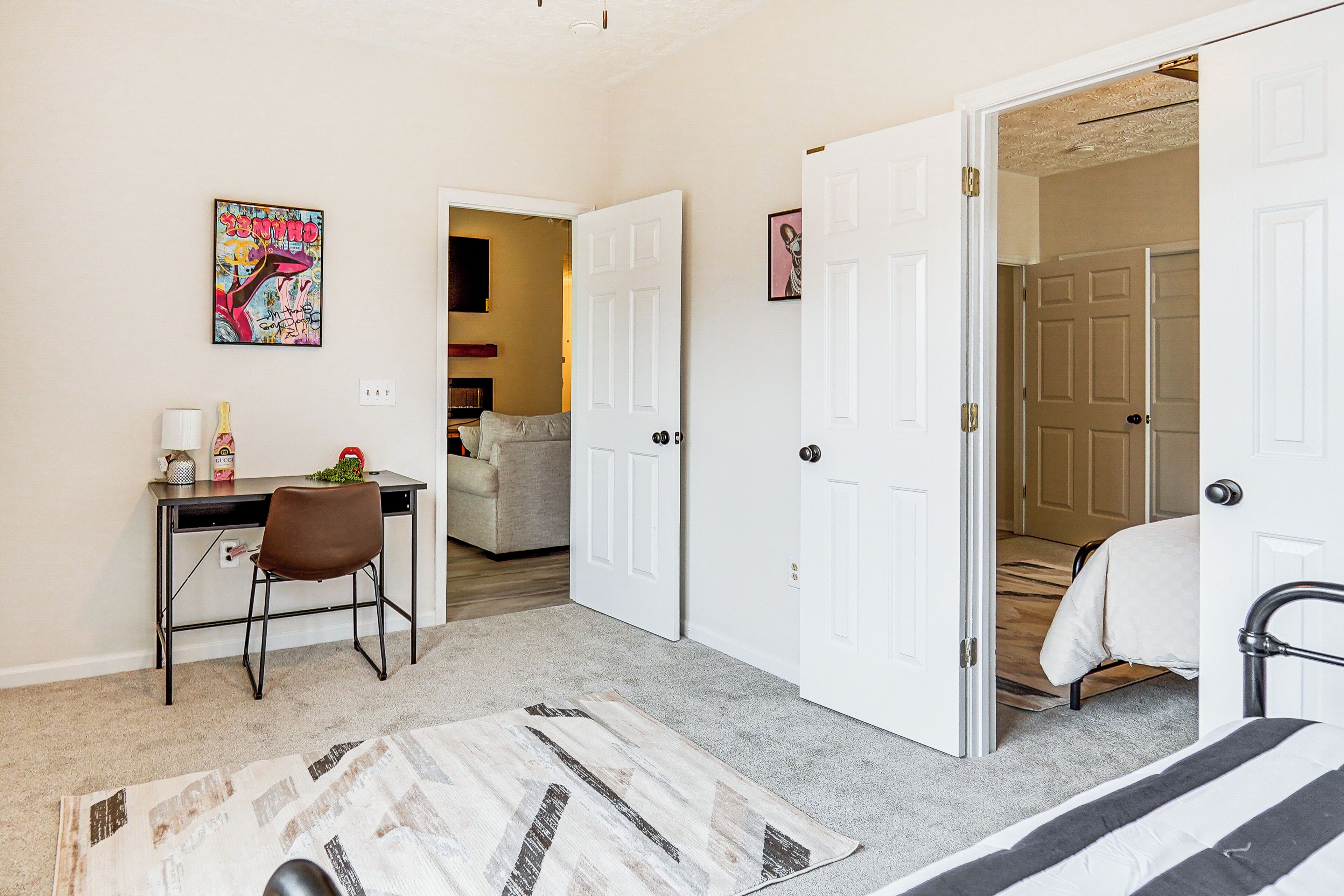 A bedroom with a bed, a desk with a chair, a door, and a hallway leading to another room.