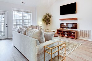 A modern living room with a sofa, a TV, a wooden TV stand, a floor lamp, and a large window. - 9 Mi to Dtwn Fayetteville: Condo w/ Pool Access! (Fayetteville)