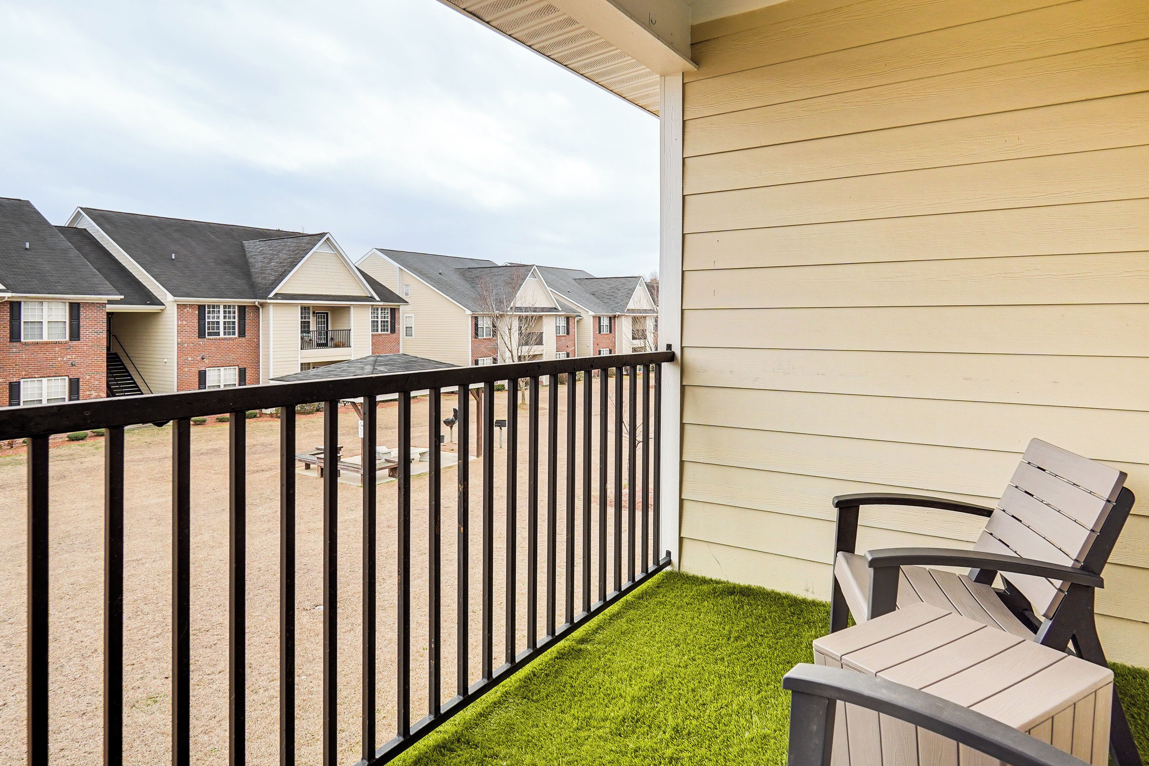 A balcony with a view of neighboring houses, a small grassy area, and a chair.
