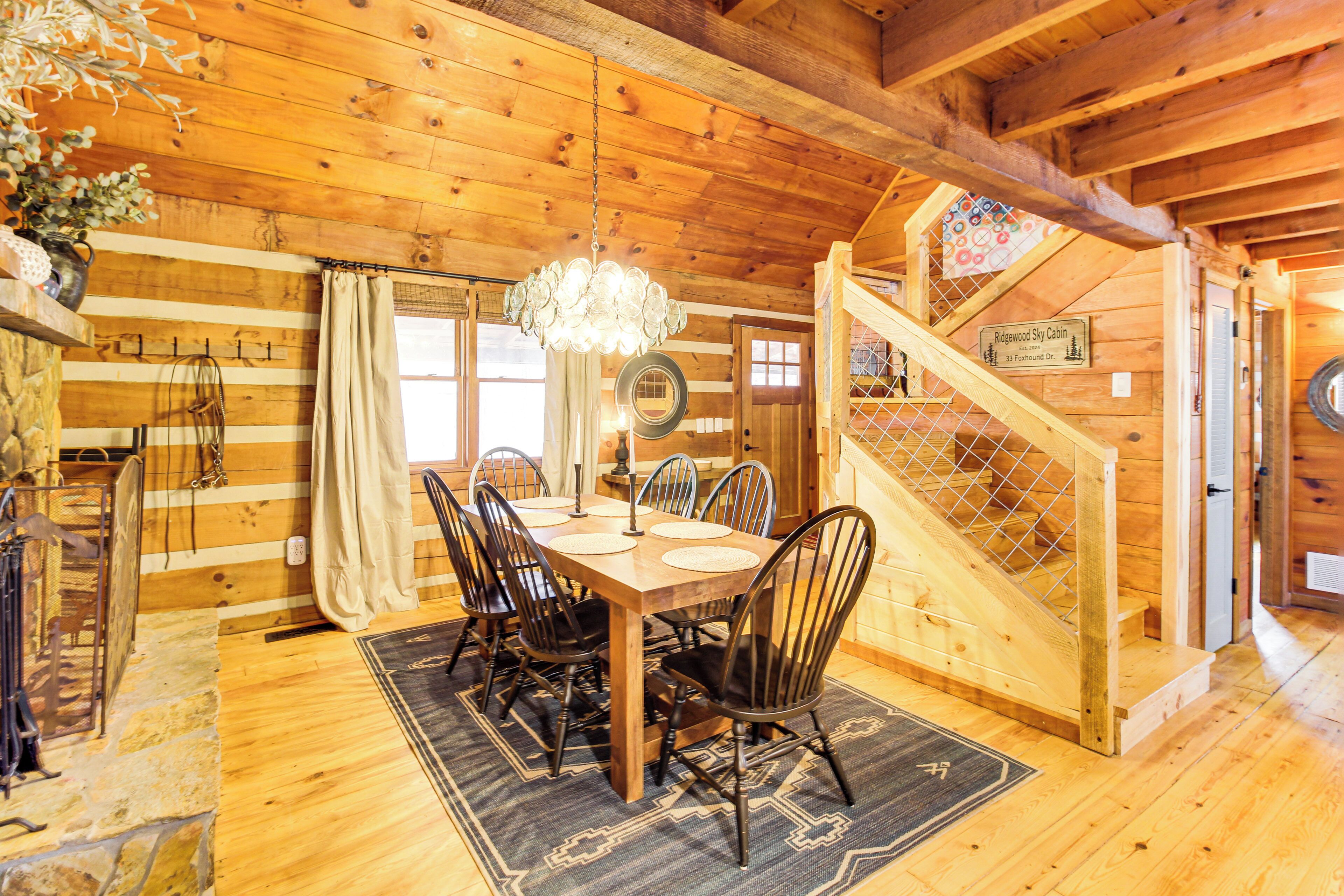 A wooden cabin interior with a dining table, chairs, a chandelier, and a staircase.