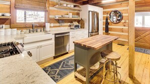 Una cocina con armarios blancos, un refrigerador de acero inoxidable, una isla de madera con taburetes y una ventana con vista a árboles.