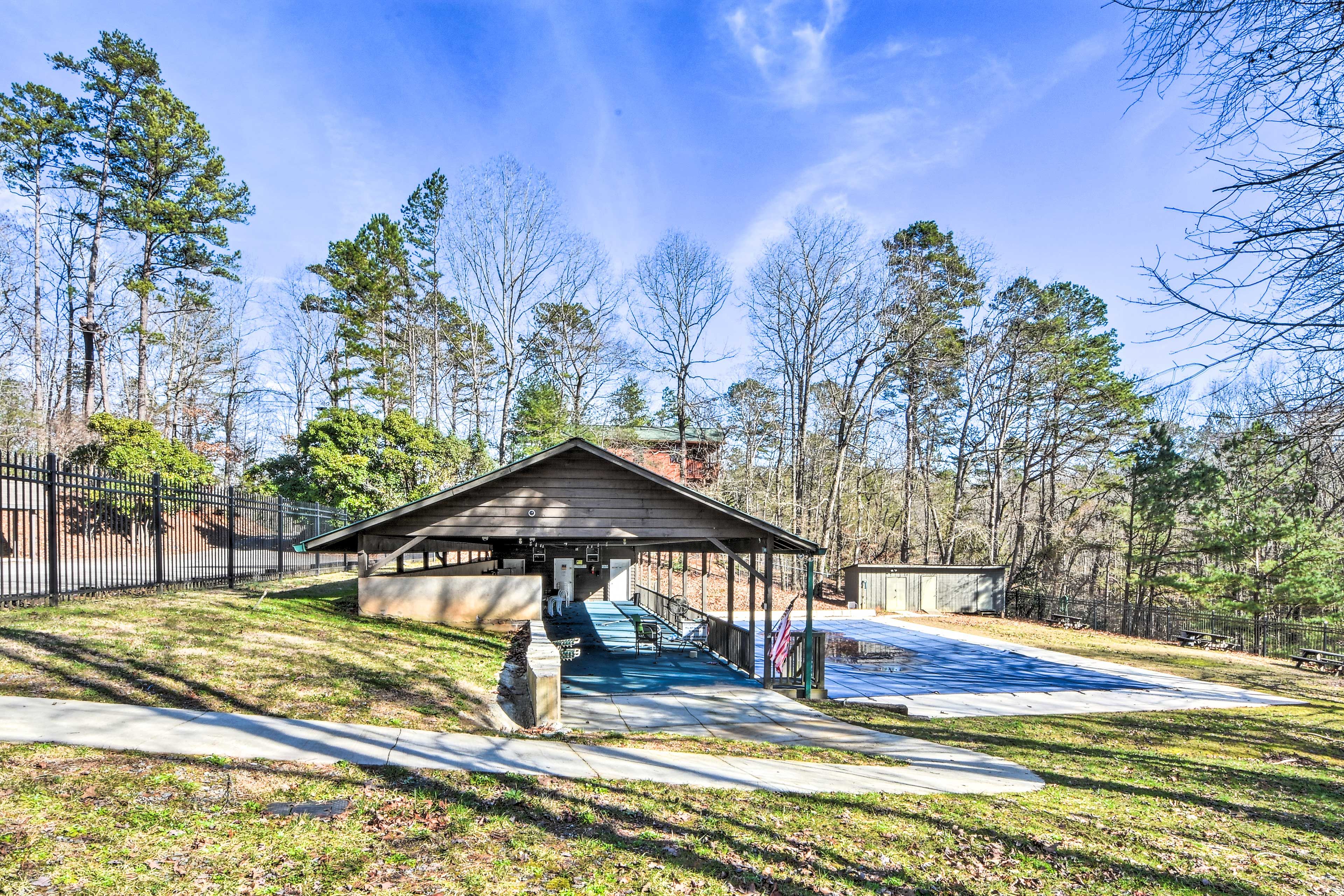 A covered pavilion with a concrete floor, surrounded by trees and a fenced area.