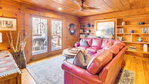 Interior acogedor de una cabaña con un sofá de cuero, estanterías de madera y un ventanal con vistas a un bosque.