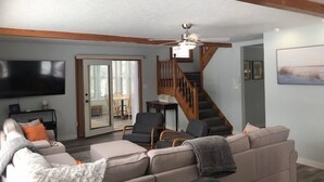 A spacious living room with a large sectional sofa, a flat-screen TV mounted on the wall, a wooden staircase, and a dining area visible through a glass door. - Peaceful, Private Lakeside Home. Sleep 16! (Pentwater)