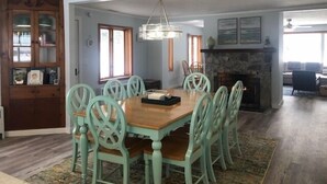 A dining area with a wooden table and green chairs, a stone fireplace, and a view of the living room. - Peaceful, Private Lakeside Home. Sleep 16! (Pentwater)