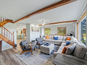 A spacious living room with a large sofa set, a wooden staircase, and a dining area visible through an open door. - Peaceful, Private Lakeside Home. Sleeps 16! Available for monthly rental! (Pentwater)