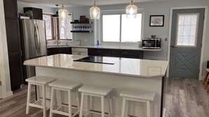 A modern kitchen with a large island, bar stools, stainless steel appliances, pendant lighting, and a view of the outdoors. - Peaceful, Private Lakeside Home. Sleep 16! (Pentwater)