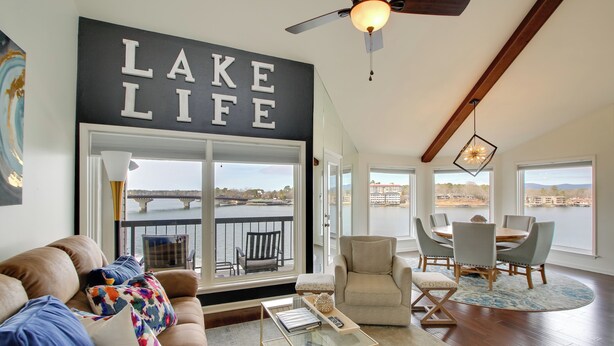 A living room with a large window overlooking a body of water, a sofa with colorful cushions, a glass coffee table, and a dining area with chairs and a table. - Million-dollar Views: Condo on Lake Hamilton! (Hot Springs)