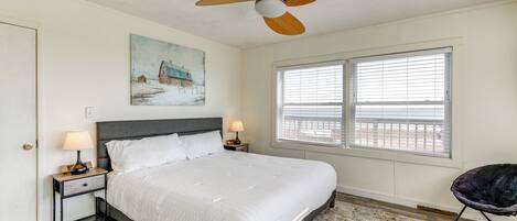A bedroom with a bed, a ceiling fan, a window with blinds, a nightstand with a lamp, and a chair.