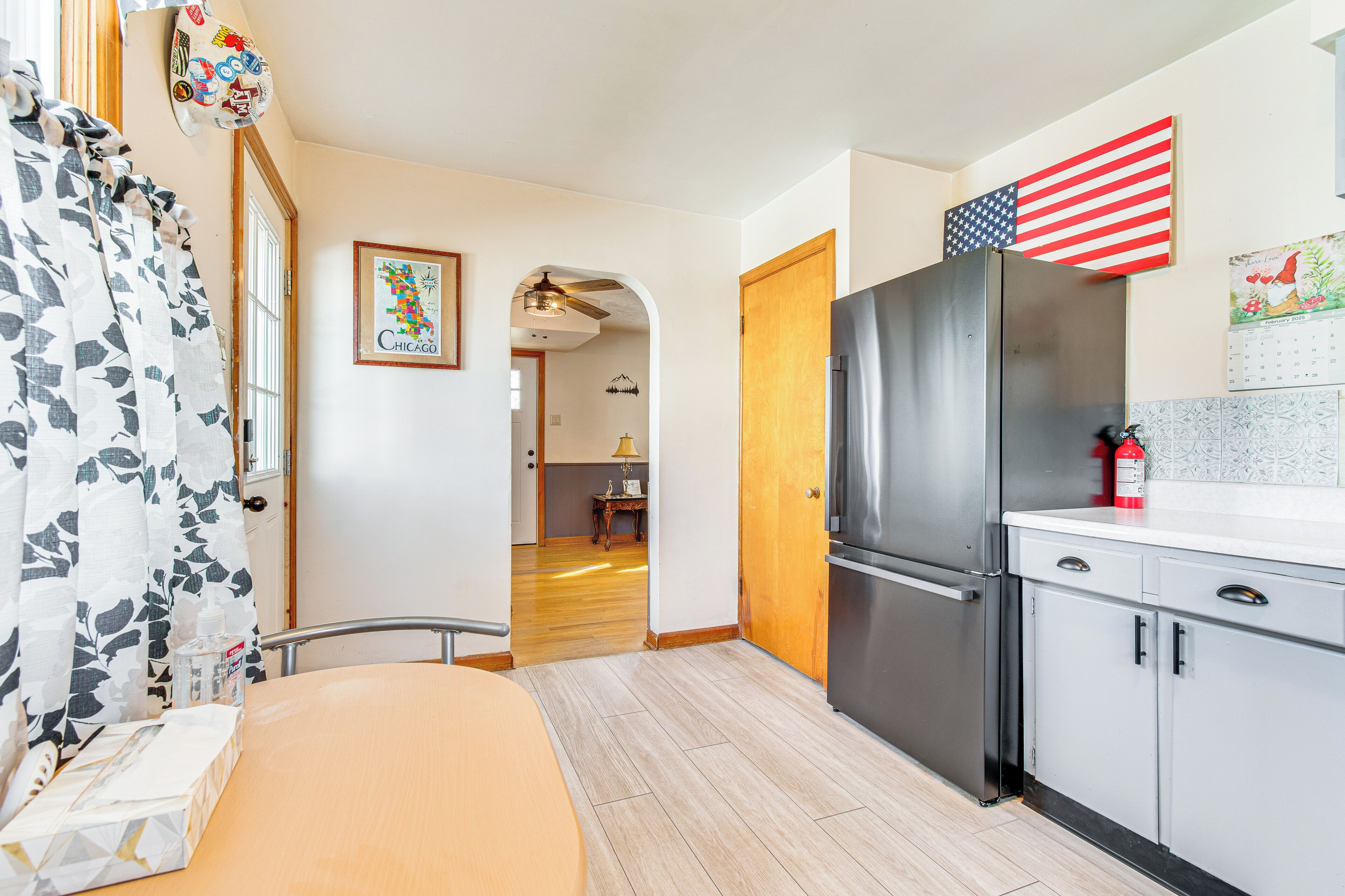 A modern kitchen with a stainless steel refrigerator, wooden cabinets, and a small table with a lamp.