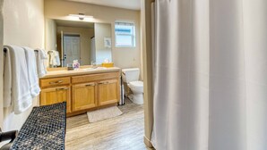 A bathroom with a wooden vanity, a toilet, a shower curtain, and a bench. - Heart of Sisters Suite (Sisters)