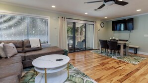 A living room with a brown leather sofa, a round coffee table, a ceiling fan, a TV mounted on the wall, and a dining area with a table and chairs. - 34 Forest Gardens: Walk to Beach, Pool, Free Bikes (Hilton Head Island)