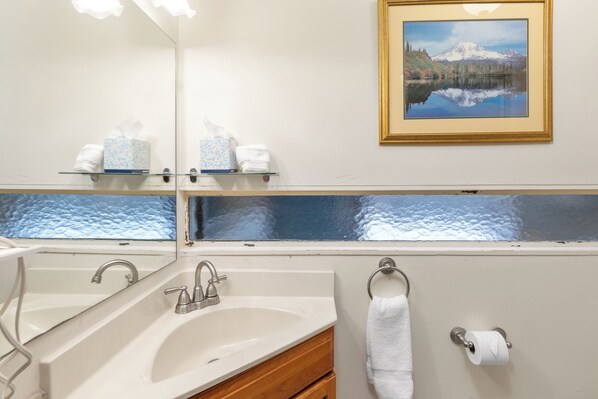 Un baño con un espejo grande, un lavabo, un perchero para toallas y un cuadro con un paisaje de montaña.