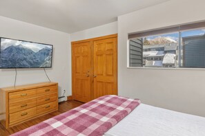 Un dormitorio con un aparador de madera, un televisor montado en la pared, una ventana con vista a un edificio y una colcha a cuadros en la cama.