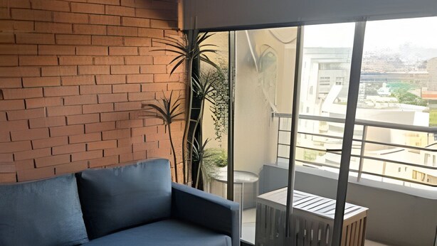 A modern living room with a grey sofa, a small round table with a plant and remotes, and a sliding glass door leading to an outdoor balcony with a view of buildings. - NR APART Bellas Artes (Santiago)