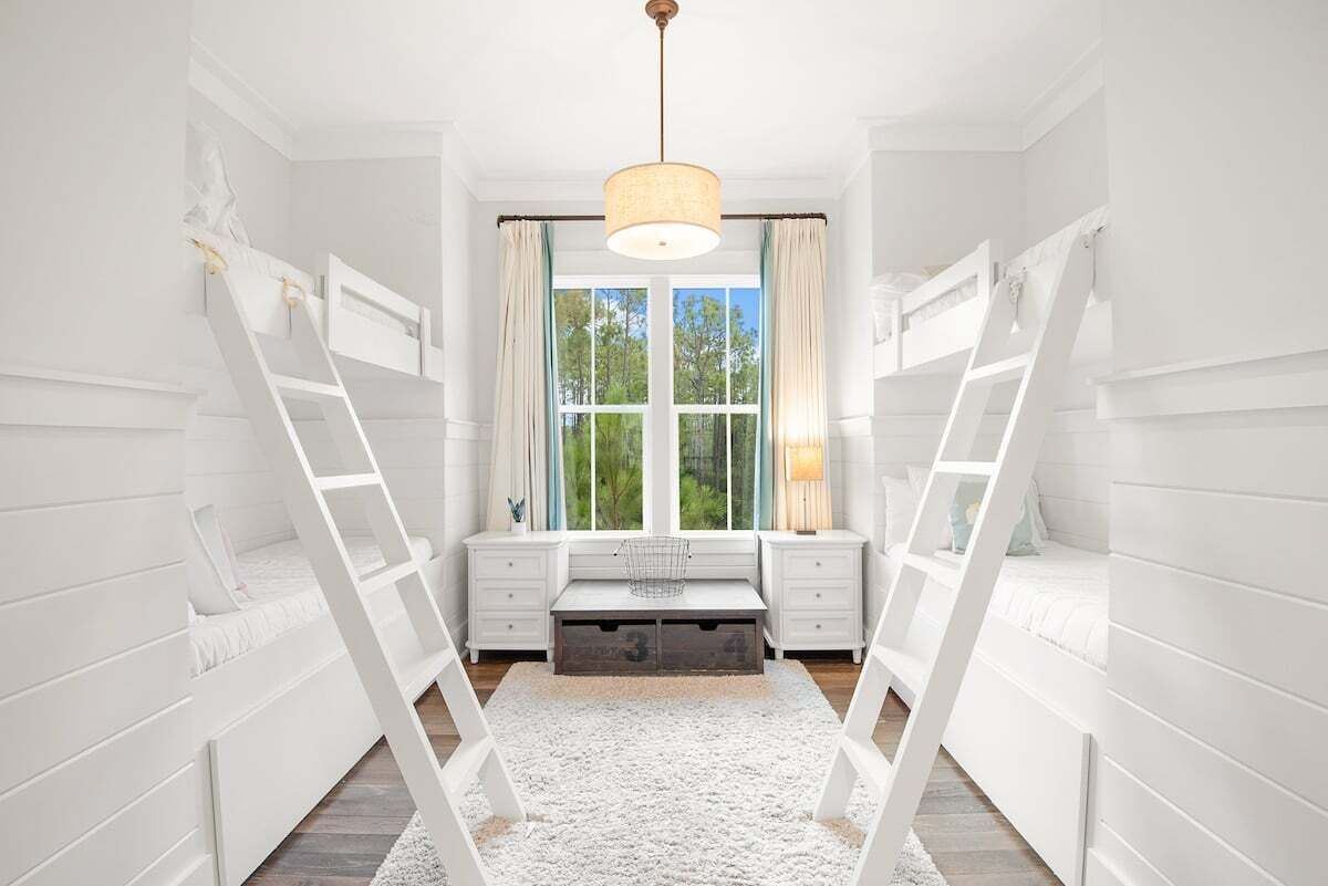 Una habitación blanca con literas, una ventana con cortinas, un escritorio y una lámpara.