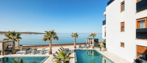 Zona de piscina con dos piscinas, palmeras, sillas de descanso y vistas al mar.