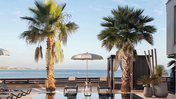 A poolside area with lounge chairs, umbrellas, and palm trees, overlooking a body of water. - Malibu Imperial Resort (Vir)