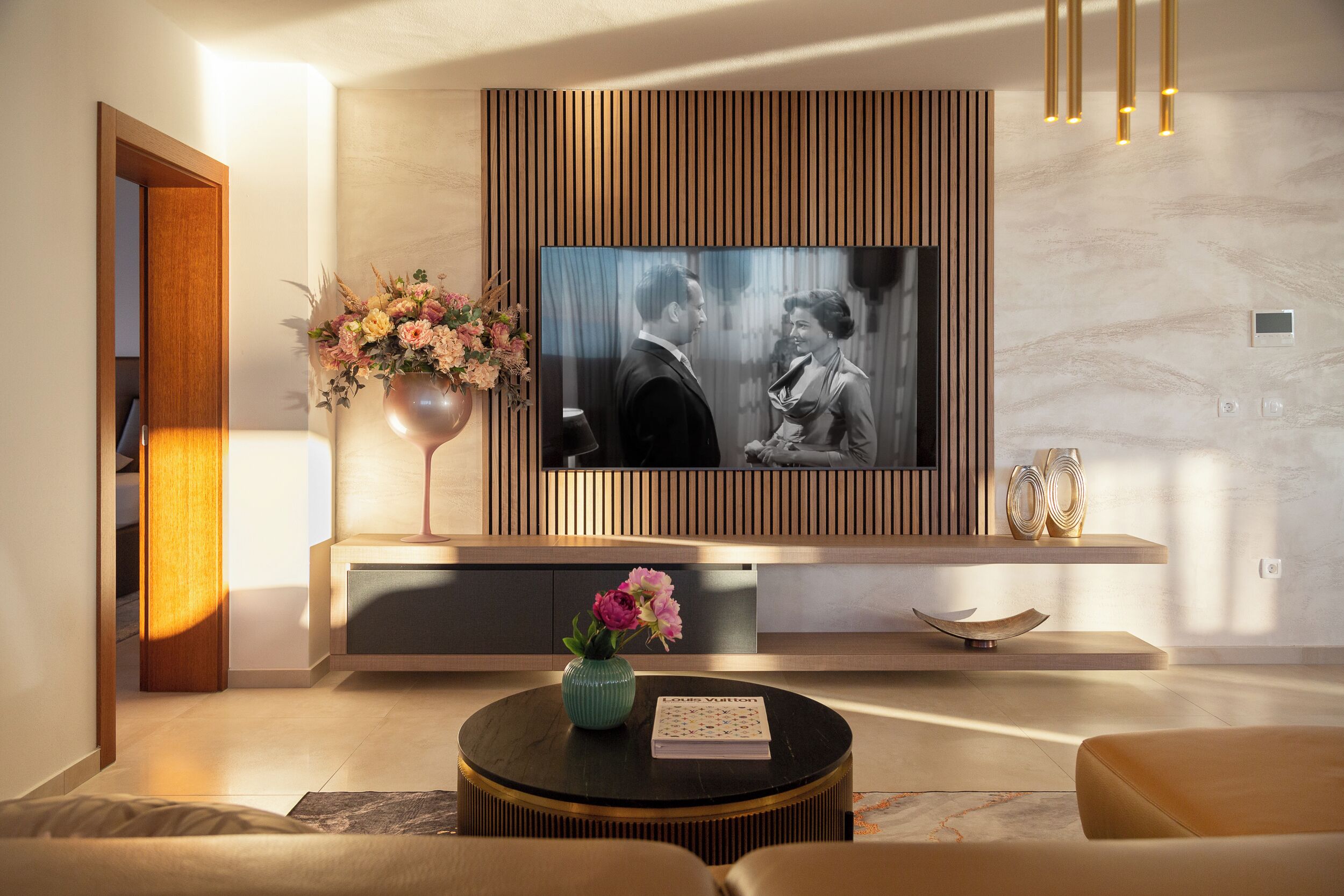 A modern living room with a flat-screen TV mounted on a wooden panel wall, a coffee table with a vase of flowers, and a sofa.