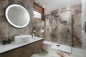 A modern bathroom with a round mirror, a white sink, a glass-enclosed shower, and a toilet.