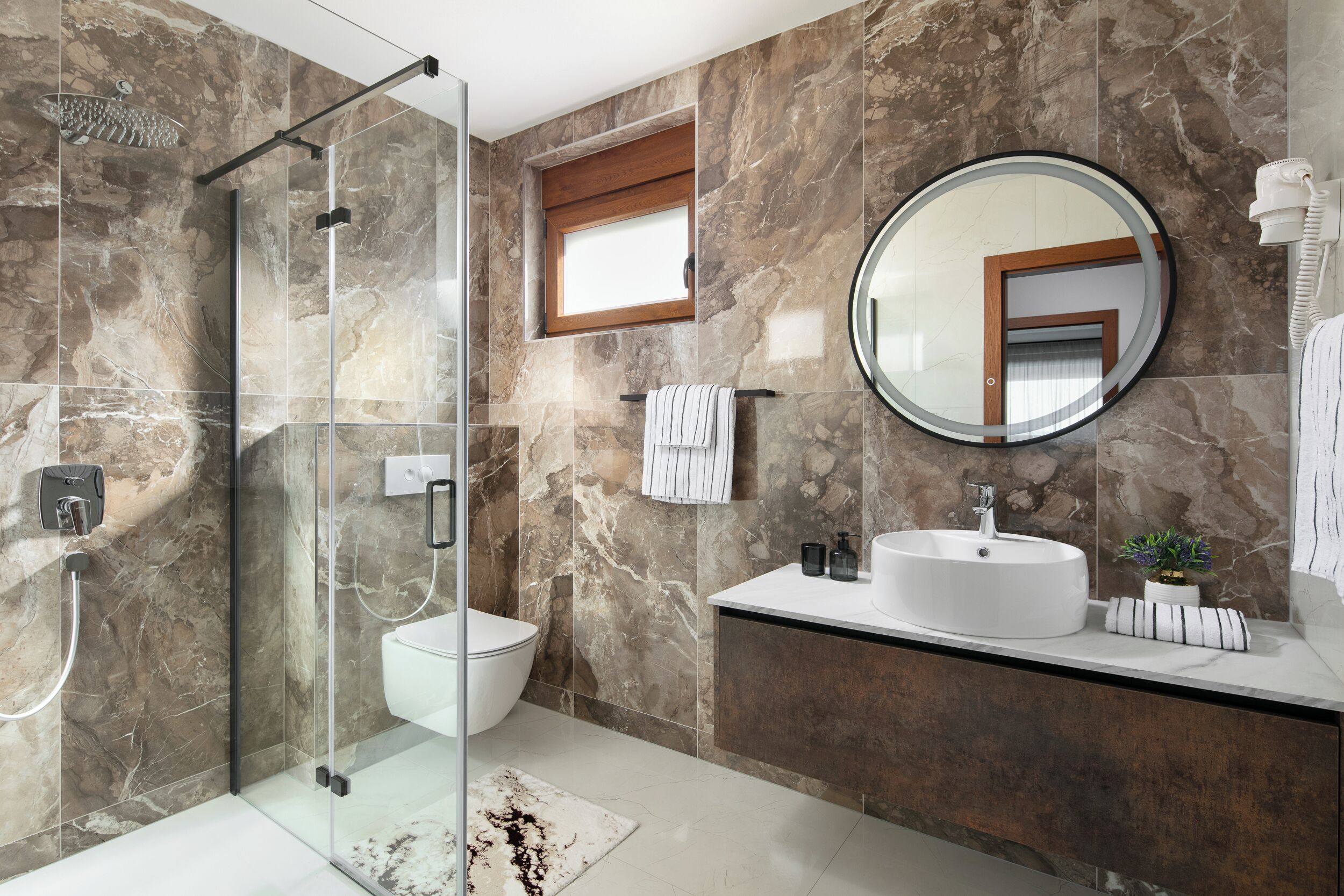 A modern bathroom with a glass-enclosed shower, a round mirror, a white sink, and a marble-tiled wall.