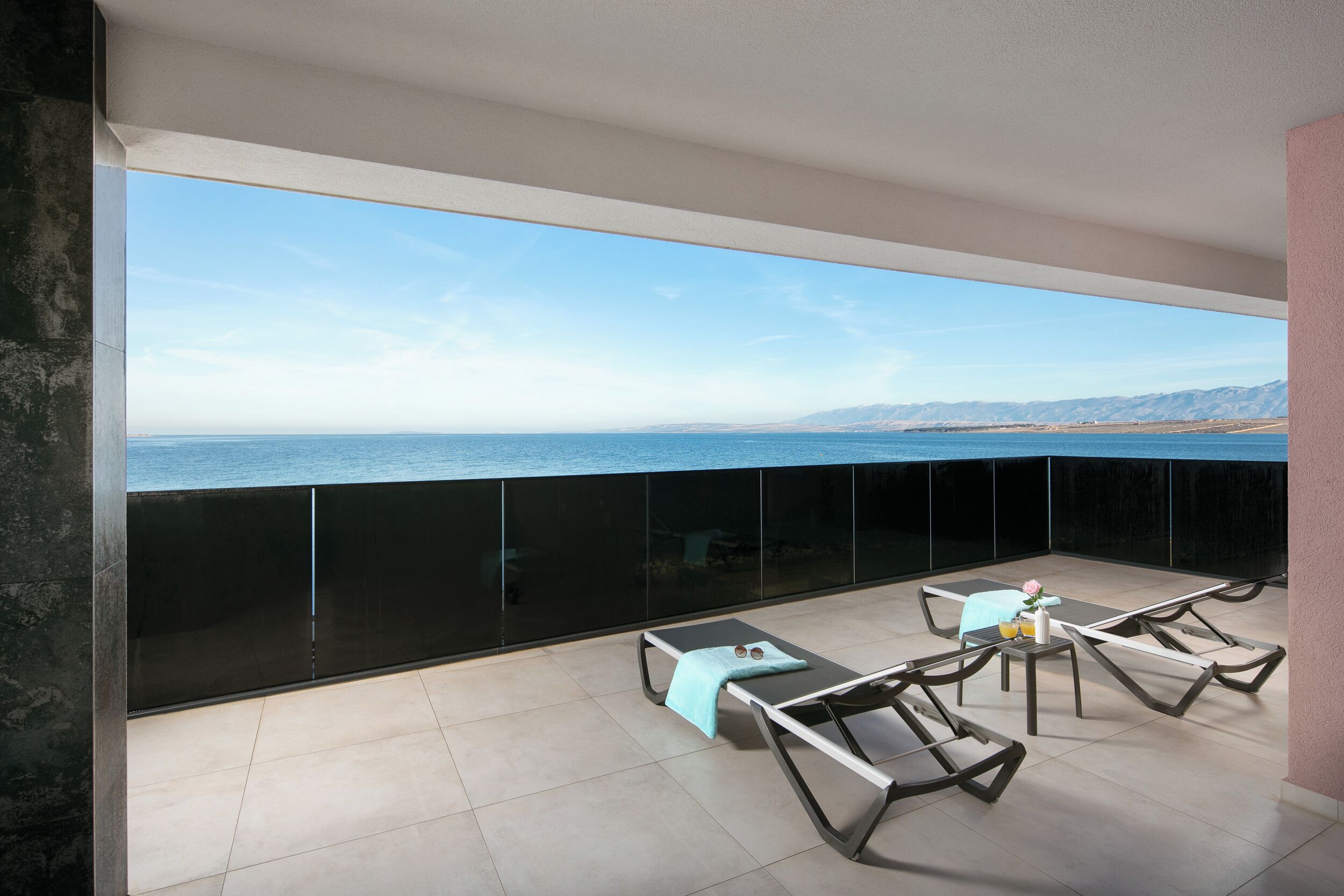 A balcony with a view of the sea and mountains, furnished with lounge chairs and a table.