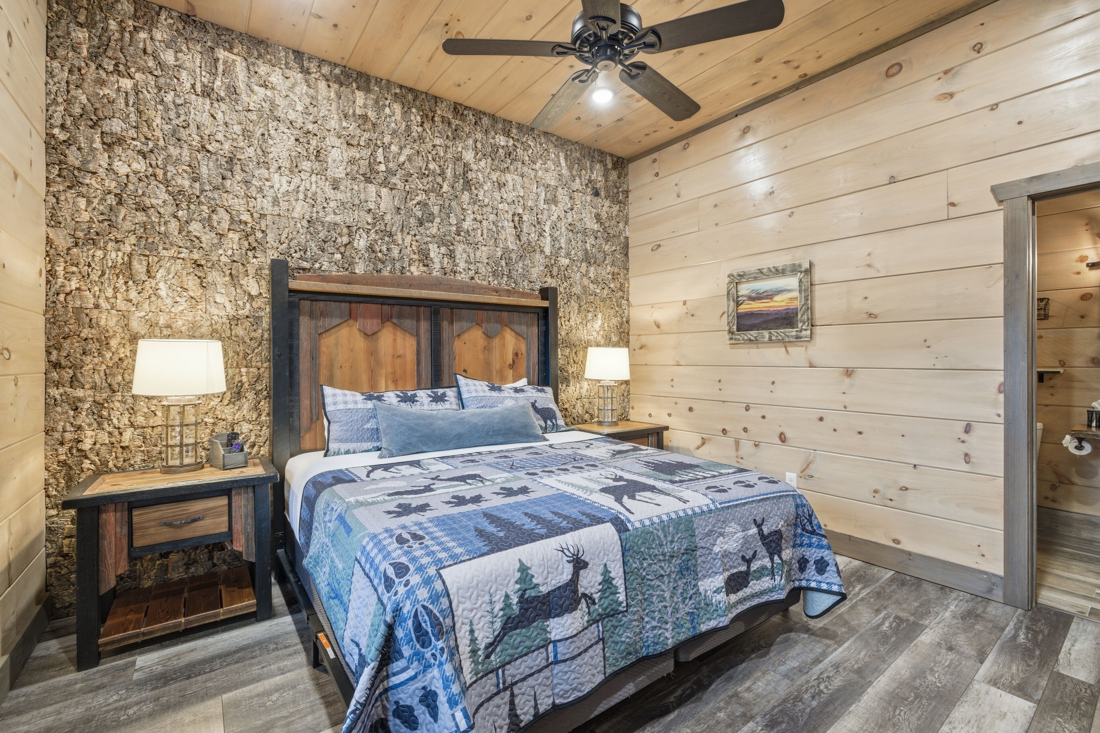 A bedroom with a wooden headboard, a bed with a patterned quilt, two bedside lamps, a stone wall, and a ceiling fan.