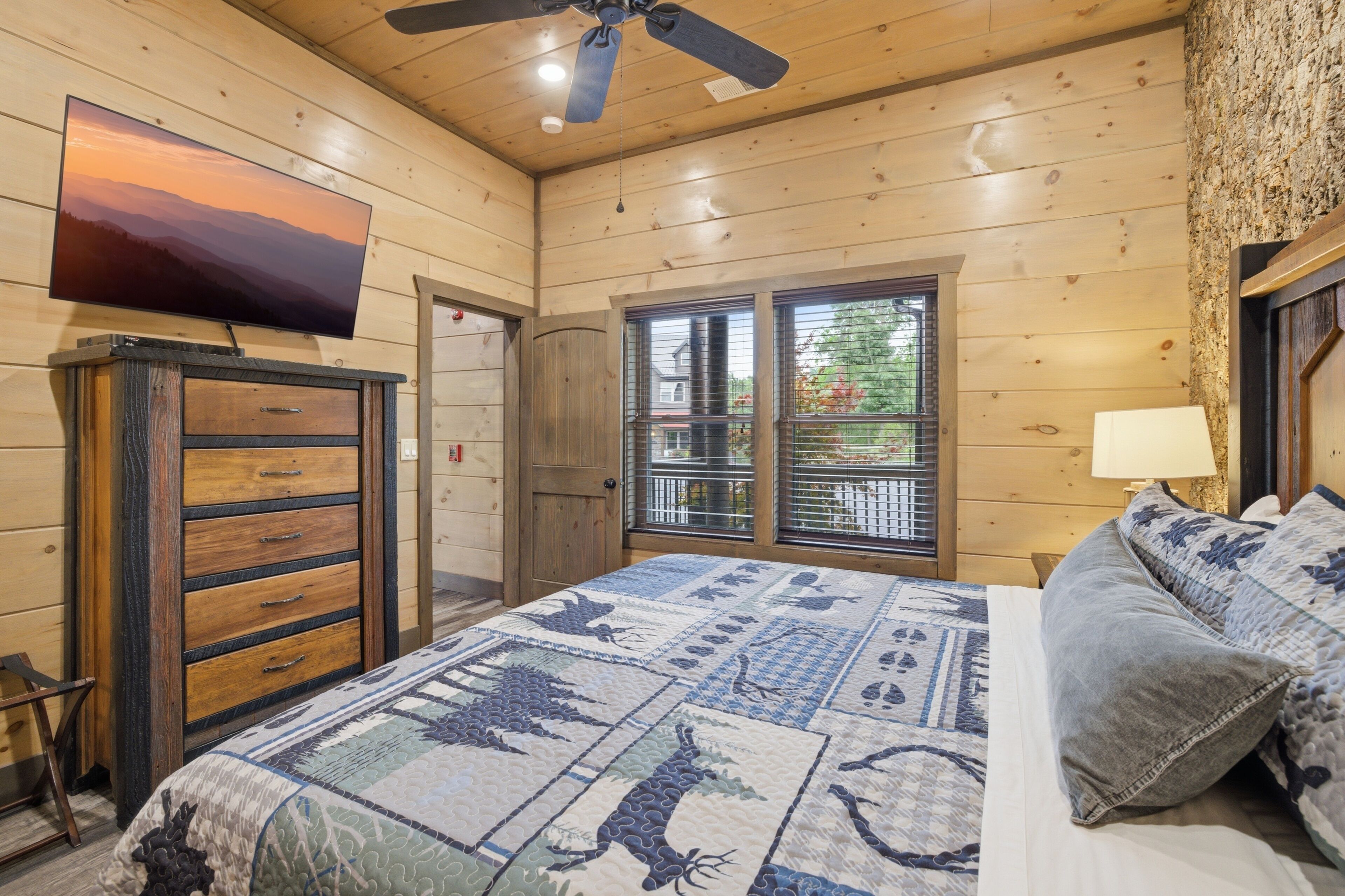 Ein Schlafzimmer einer Holzhütte mit einem Bett, einer Kommode, einem Fernseher und einem Fenster mit Blick nach draußen.