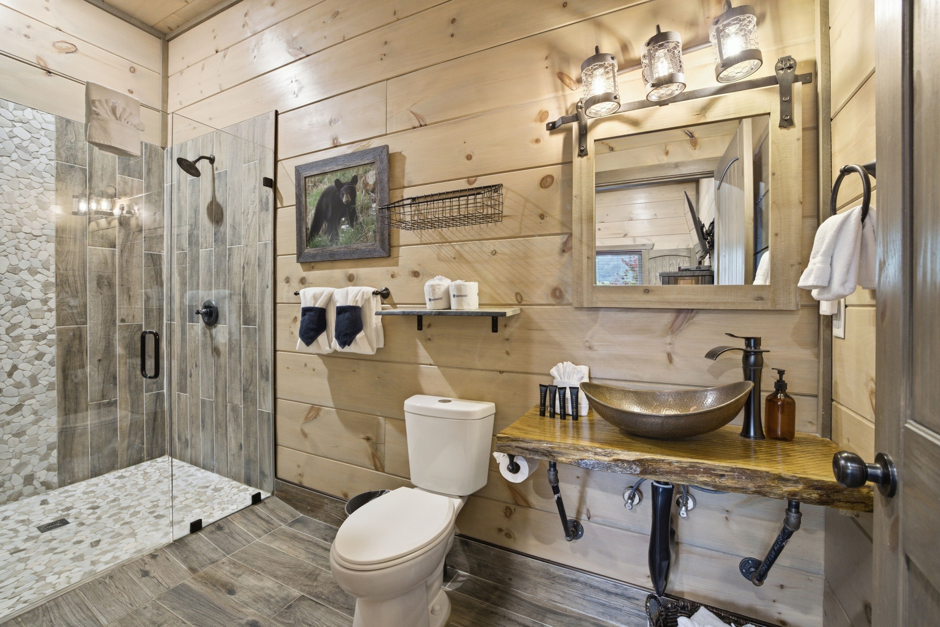 A wooden-paneled bathroom with a glass shower enclosure, a toilet, a sink with a vessel basin, and a mirror with lights.