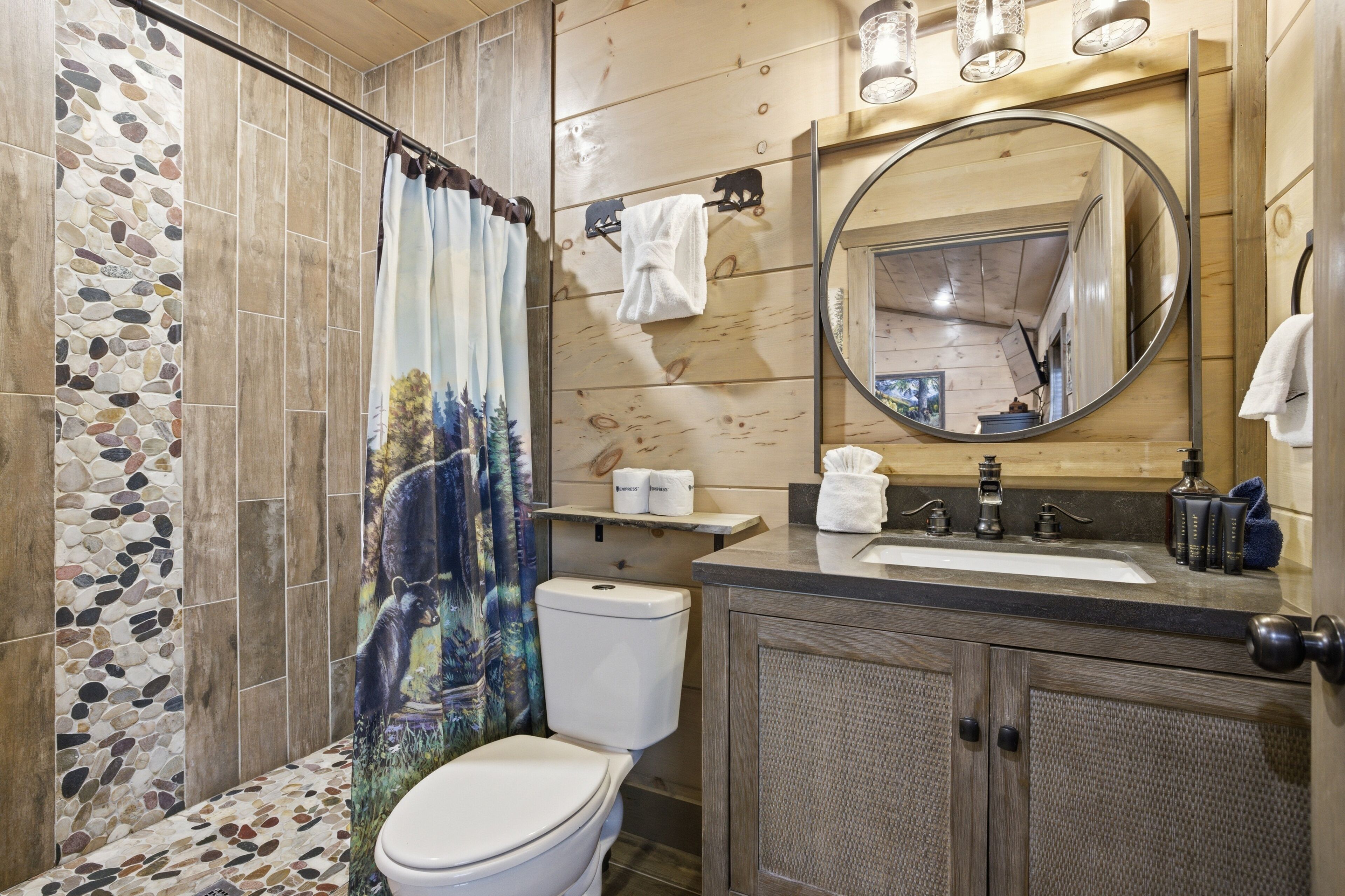 Une salle de bain avec un mur recouvert d’une mosaïque de pierre, des toilettes, un rideau de douche orné d’un motif d’ours, un meuble-lavabo en bois avec un miroir et un lavabo avec des articles de toilette.