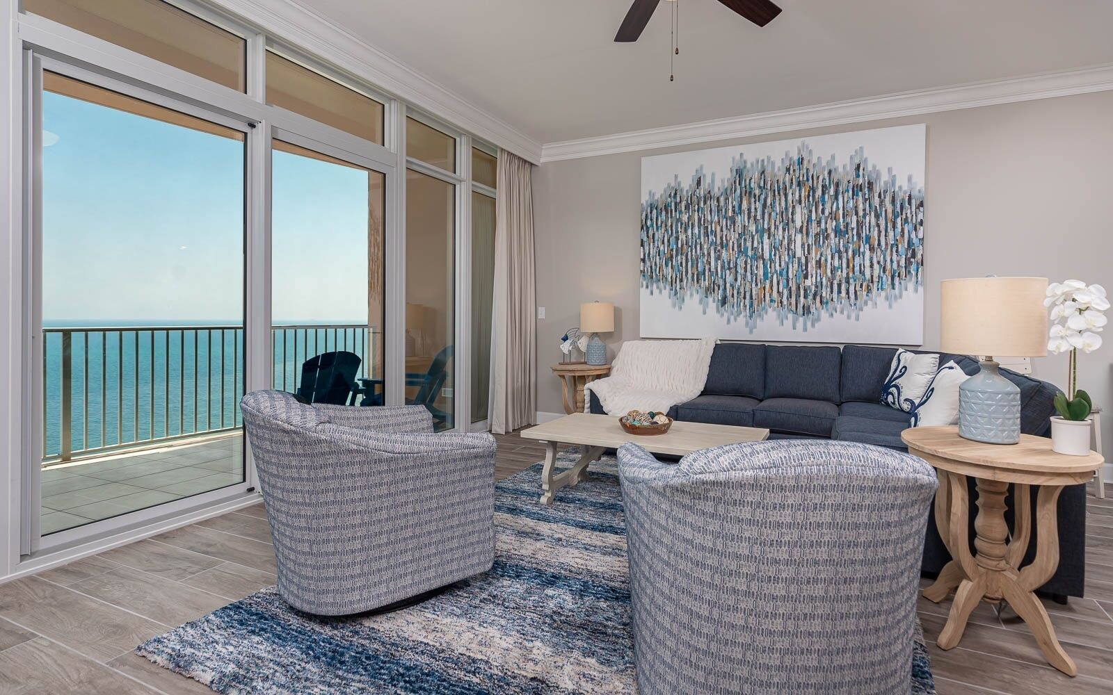 A spacious living room with a large sliding glass door leading to a balcony, a grey sofa, two armchairs, a coffee table, and a decorative abstract painting on the wall.