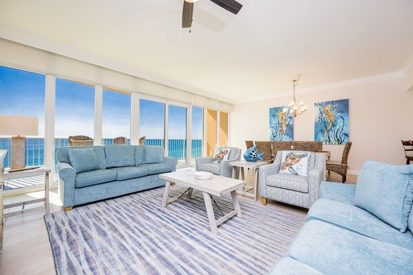 A spacious living room with a large window, a blue sofa, a coffee table, and a chandelier. - Phoenix Gulf Towers 1006 2 Bedroom Condo (Orange Beach)