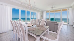 A dining area with a glass table, white chairs, and a chandelier. It overlooks a beach with chairs and tables.