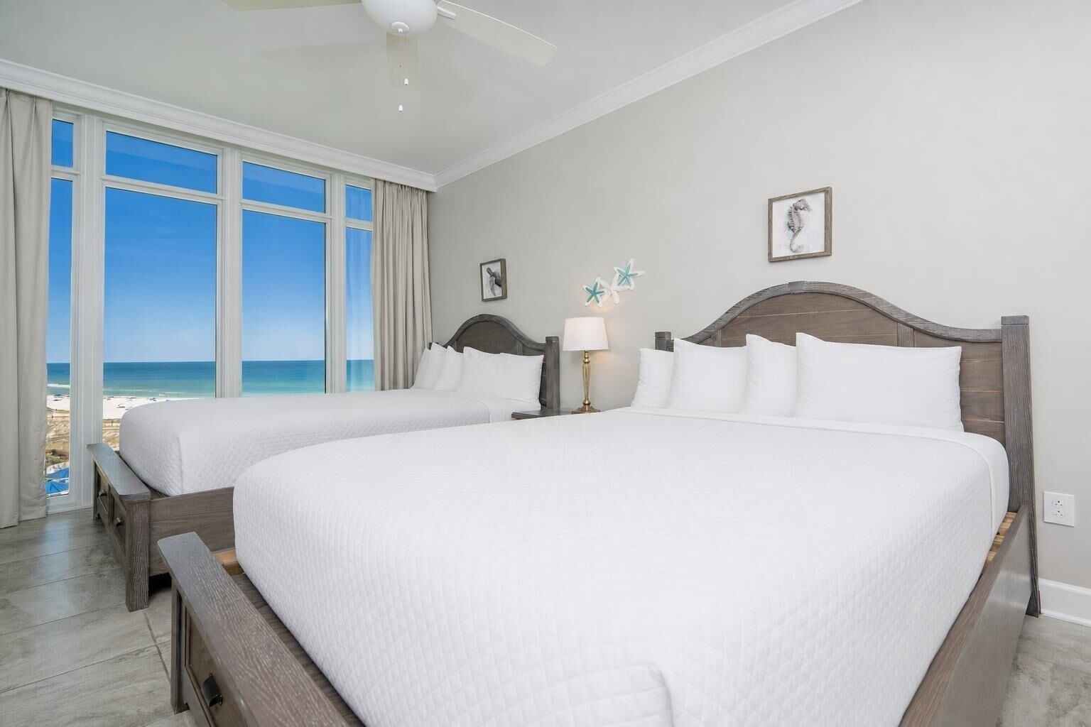 A hotel room with two beds, a large window with a beach view, a ceiling fan, and a wall-mounted lamp.