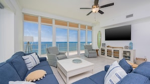 A spacious living room with a large window, a ceiling fan, a flat-screen TV, and a white coffee table.
