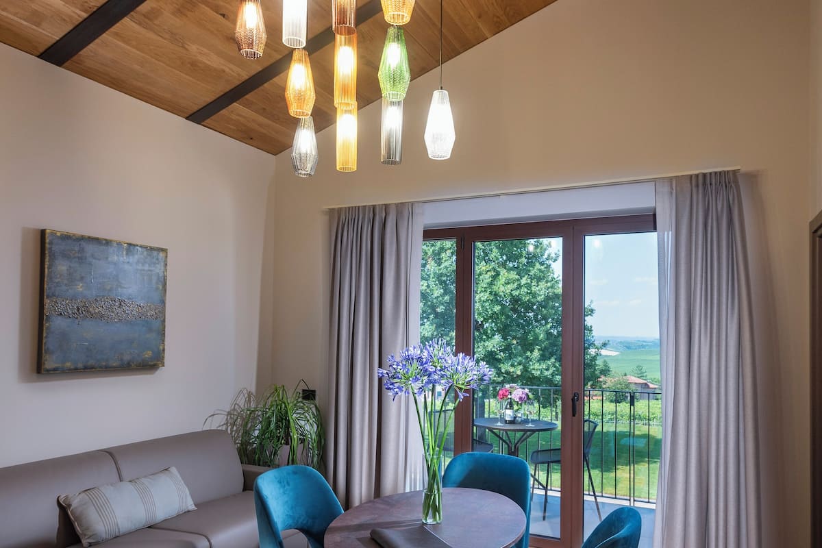 A modern living room with a wooden ceiling, a sofa, a dining table with chairs, and a view of the outdoors through large windows.
