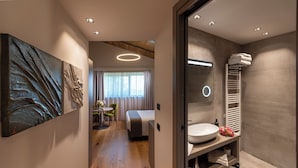 Une chambre d’hôtel moderne avec un grand tableau abstrait, une table ronde avec des chaises, un lit, une salle de bain avec un lavabo et un porte-serviettes, et une vue sur le couloir.