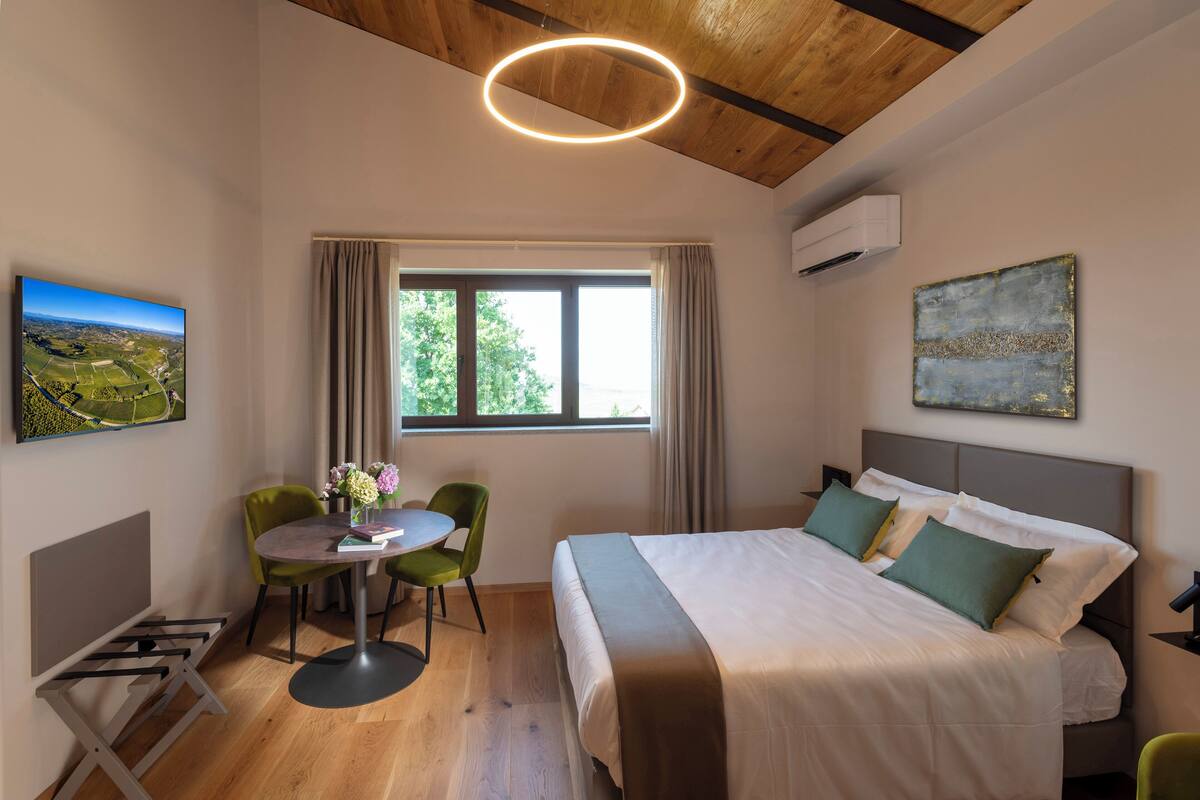 A hotel room with a bed, a desk with a chair, a window with curtains, and artwork on the walls.