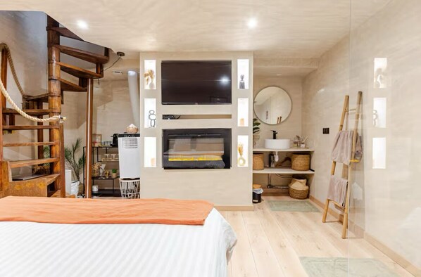 A bedroom with a bed, a TV mounted on the wall, a wooden ladder, and a small table with a lamp.
