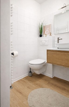A modern bathroom with white tiles, a wooden floor, a toilet, a sink with a mirror, and a small plant on the countertop.