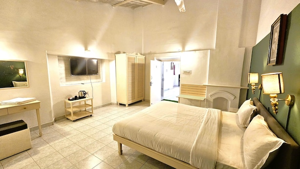 Quarto de hotel com uma cama grande, uma escrivaninha com cadeira, um guarda-roupa, uma televisão e um banheiro visível por uma porta aberta.