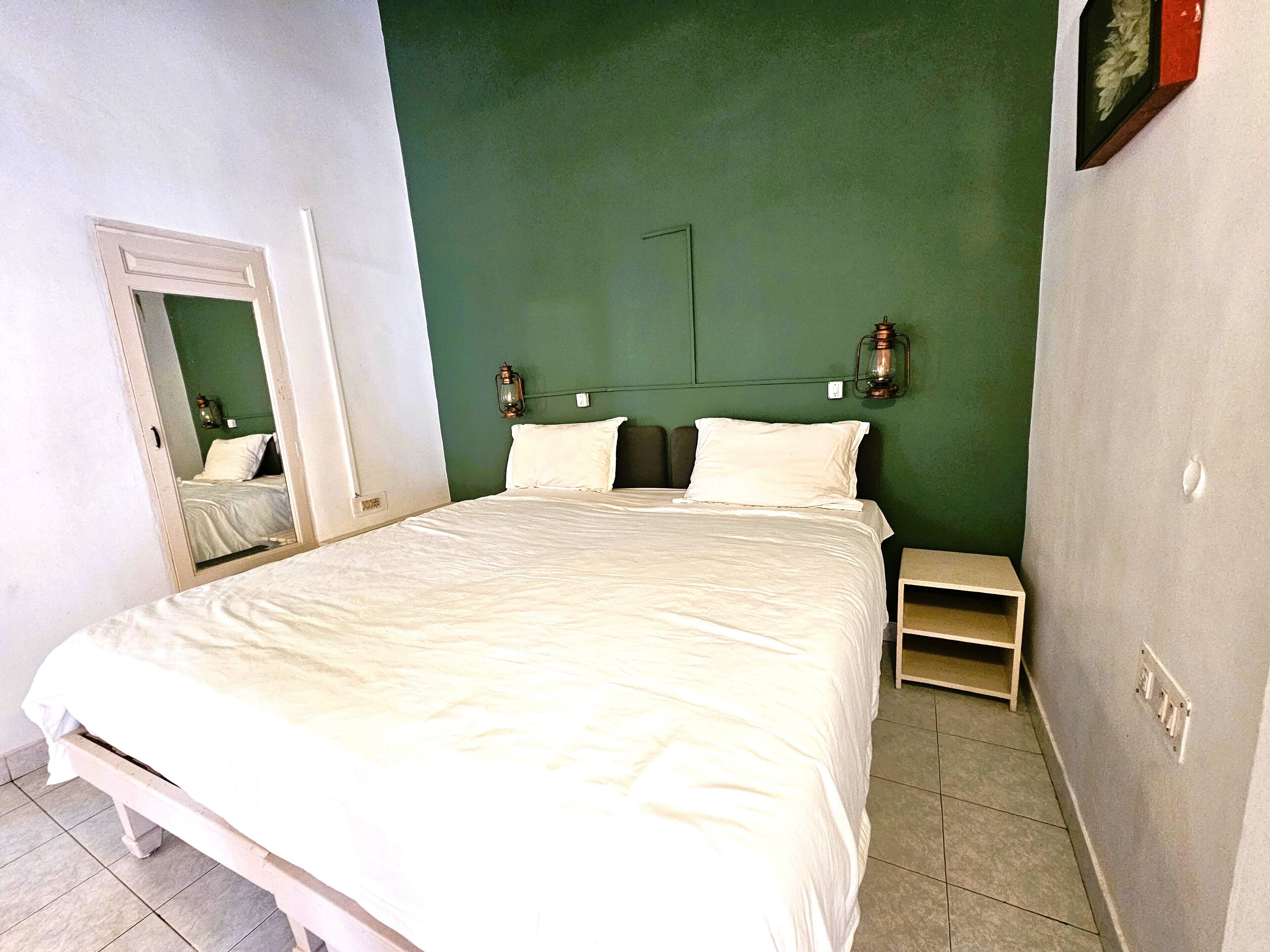 A neatly made bed in a room with green walls, a small bedside table, and a mirror on the left wall.