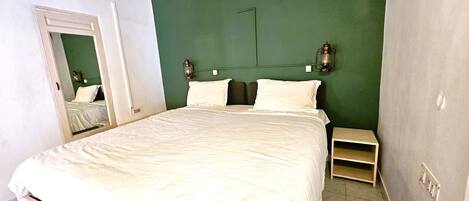 A neatly made bed in a room with green walls, a small bedside table, and a mirror on the left wall.