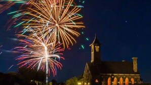 Een nachtelijke scène met vuurwerk boven een wateroppervlak, dat een gebouw met een klokkentoren weerspiegelt.