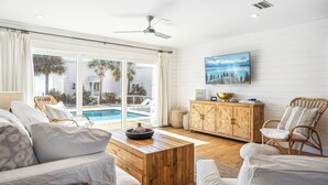 A spacious living room with a wooden coffee table, white sofas, and wicker chairs. A large window offers a view of a pool and palm trees. - Private Beach Access, Private Pool/Hot Tub, Beach Service, and Pet Friendly! (Santa Rosa Beach)